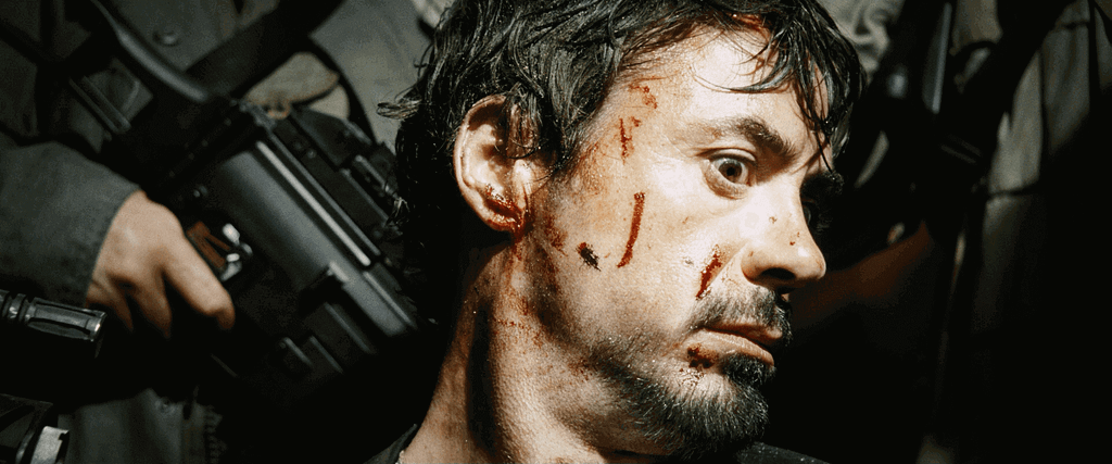 Tony Stark, bruised and bloodied, held captive by armed men in a tense scene from Iron Man.