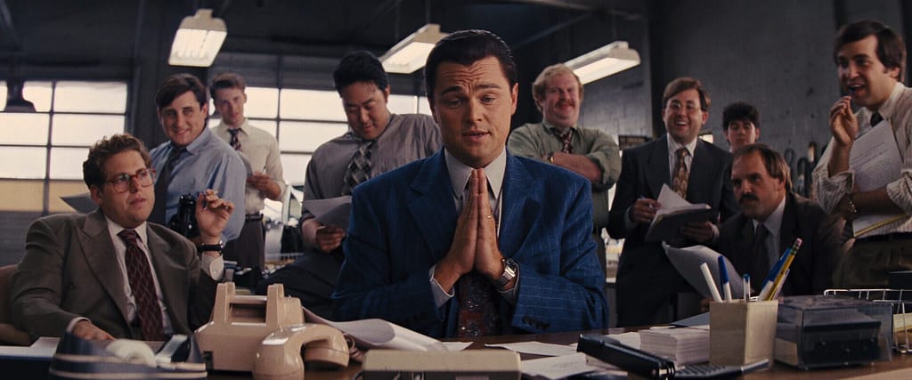 Jordan Belfort sits confidently at a desk surrounded by enthusiastic coworkers in a chaotic office scene.