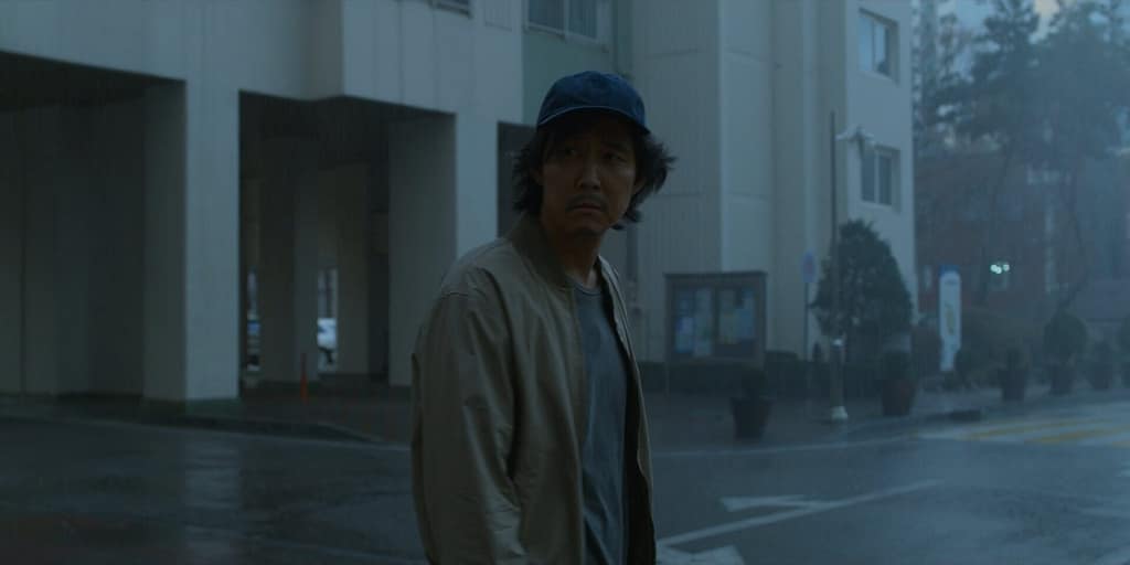Gi-hun from Squid Game standing in the rain outside an apartment complex, reflecting emotional exhaustion, guilt, and post-traumatic grief.