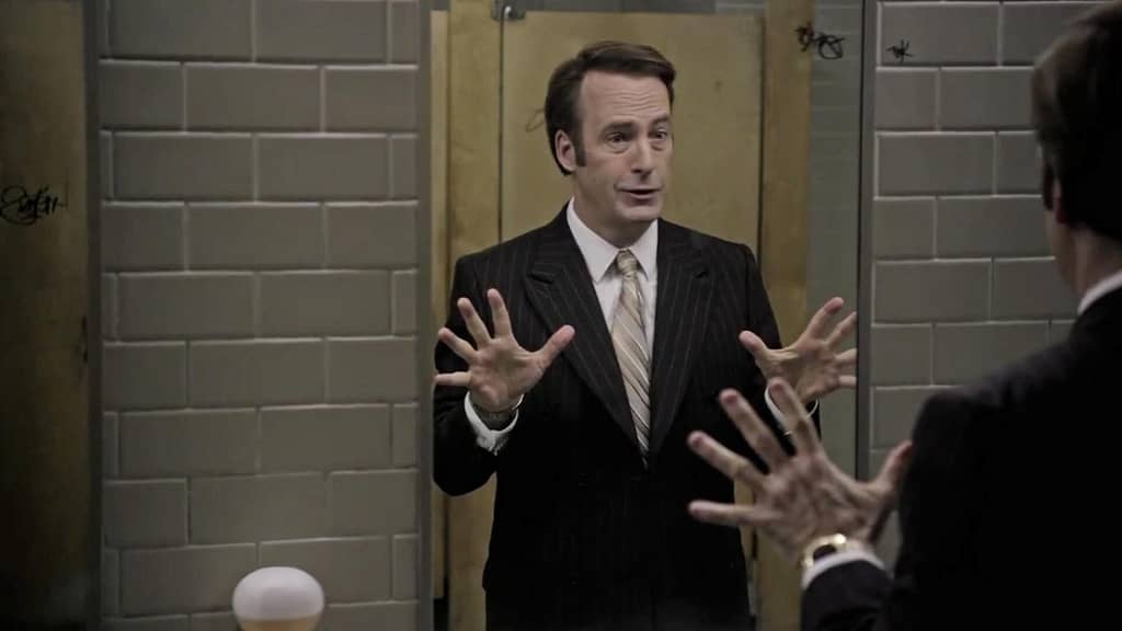 Man in a suit practicing confident body language in front of a bathroom mirror, using open hand gestures.