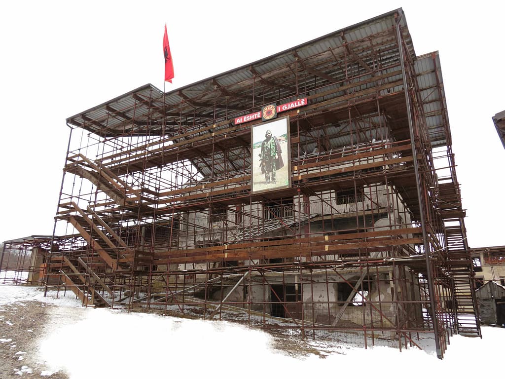 Partially destroyed building wrapped in scaffolding with Albanian flag and wartime portrait.