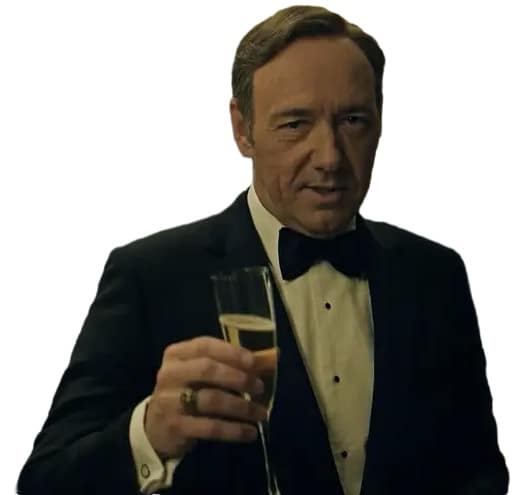 Frank Underwood in a tuxedo holding a champagne glass at a formal event with guests and blue balloons in the background.
