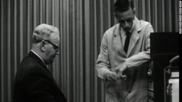 Black-and-white photo of a researcher in a lab coat standing beside an older man during the Milgram obedience study.