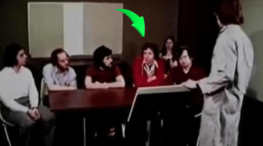 1970s experiment scene with a group seated around a table, one man in red sweater highlighted