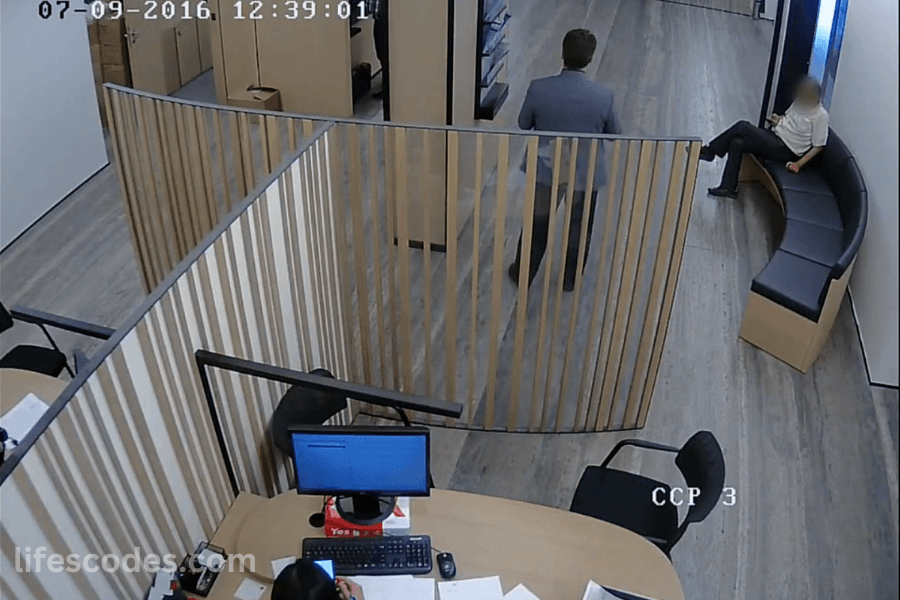Man walking through office cubicles as surveillance captures front desk employee and waiting visitor