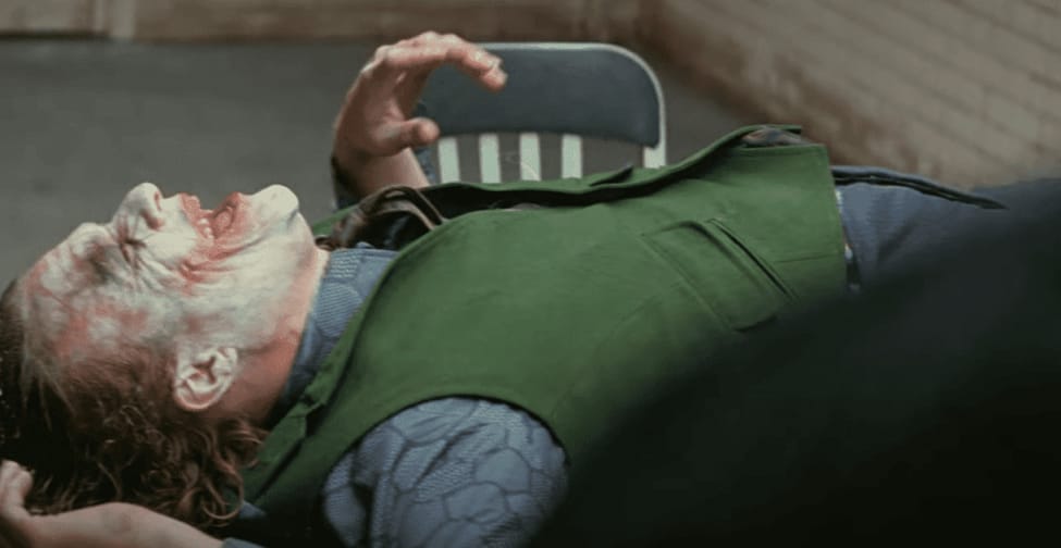 Joker lies on a table laughing during interrogation, makeup smeared, shirt wrinkled.