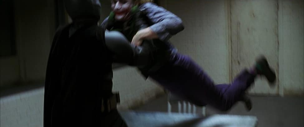 Batman slams Joker against a table in a dimly lit interrogation room.